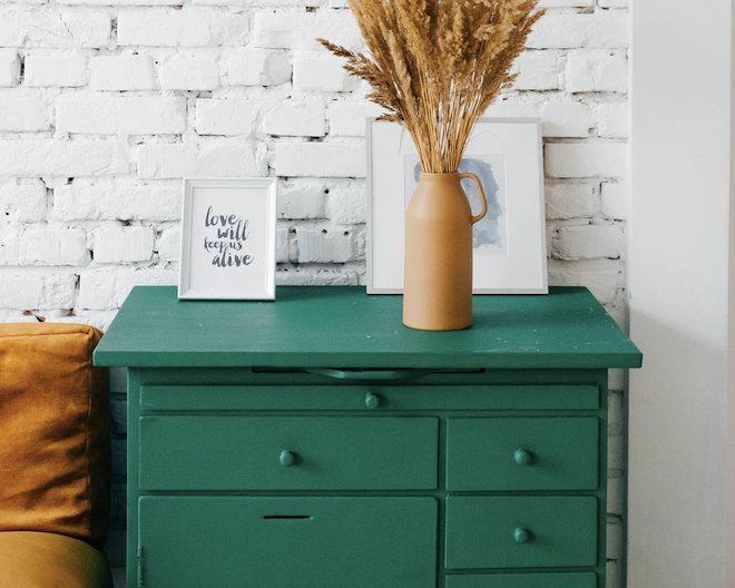 A green dresser with a decorative vase, dried wheat stalks, and a framed quote, against a white brick wall.