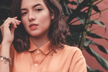 A young woman with mid-length wavy hair wearing an orange shirt with a pocket on the left side, adorned with a bracelet with charms on the left wrist, standing in front of a background of green plants.